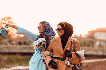 Couple woman one wearing a hijab and a modern yet traditional dress, and the other in a blue dress and scarf, walking together through the city at sunset. One carries a bouquet and bread, while the other holds a blue handbag, symbolizing a beautiful fusion of cultures, friendship, and serene urban charmの写真素材