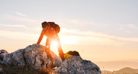 Two hikers, couple in outdoor gear climb rocky terrain with the sun rising behind them.Climbers reaching the mountain peak at sunrise.の写真素材