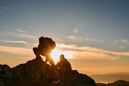 Two hikers, couple in outdoor gear climb rocky terrain with the sun rising behind them.Climbers reaching the mountain peak at sunrise.の写真素材