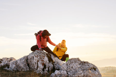 Two hikers, couple in outdoor gear climb rocky terrain with the sun rising behind them.Climbers reaching the mountain peak at sunrise.の写真素材
