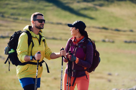 Couple hikers enjoy a scenic mountain trek in the sunshine.A man and a woman with backpacks and trekking poles walk through a green mountain landscape, smiling under the sunの写真素材