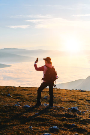A female hiker captures the sunset from the mountain peak for social media.A woman hiker enjoys the...