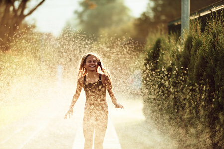 In the backyard of house, a young girl bursts with laughter and joy as she gleefully runs through a water sprinkler on a sunny summer dayの写真素材