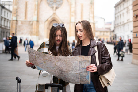 Two female tourists standing with luggage while reading a map and planning their route in the city.の写真素材