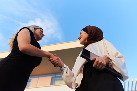 In a real-life scene, a Muslim woman in a hijab, who is a real estate business owner, shakes hands with a blonde woman after successfully selling a propertyの写真素材