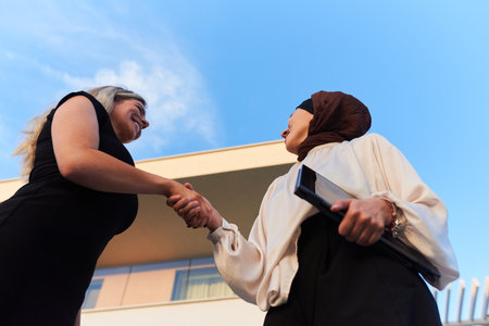 In a real-life scene, a Muslim woman in a hijab, who is a real estate business owner, shakes hands with a blonde woman after successfully selling a propertyの写真素材