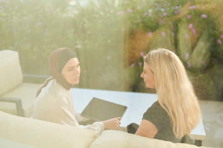 A hijab-wearing woman, a successful real estate business owner, gracefully sells a house to a blonde customer, both sitting in front of the property.の写真素材