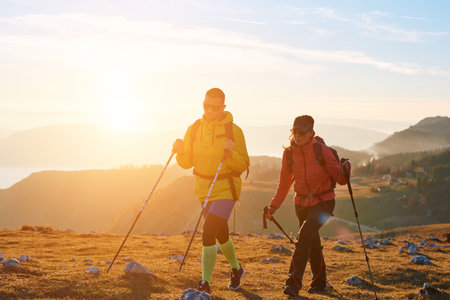 Man and woman hiking with trekking poles on mountain during golden hour, healthy active lifestyle...