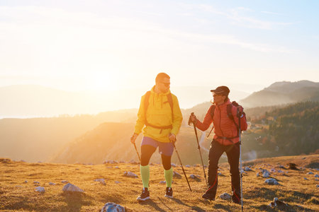 Man and woman hiking with trekking poles on mountain during golden hour, healthy active lifestyle conceptの写真素材
