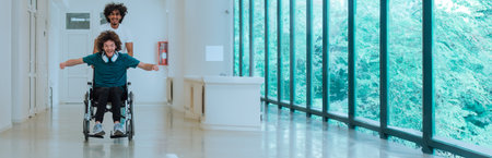 Two young friends enjoying playful wheelchair ride in modern hallway with large windows and natural...