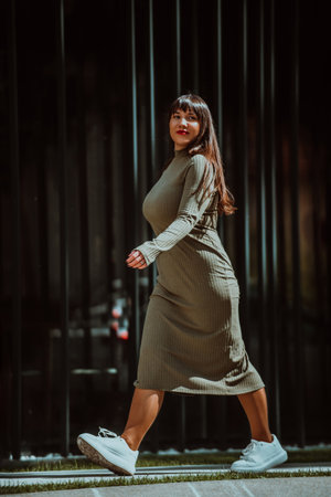 A confident and stylish plus-size woman walking by a modern building in an olive green dress, radiating beauty, empowerment, and self-love.の写真素材