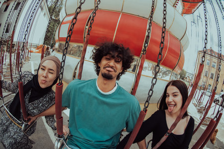 African-American man accompanied by two woman friends, one wearing a hijab, enjoys a fun-filled ride on the carousel in the amusement park, creating a vibrant and memorable moment of unity, diversity, and joy.の写真素材