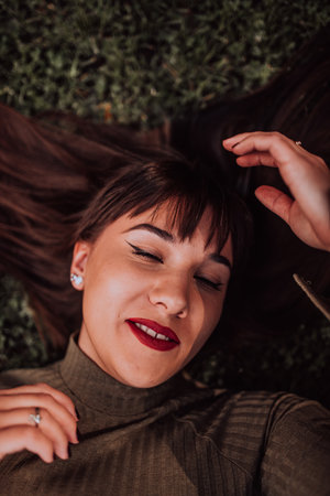 A young woman is lying on the grass, showing off her beautiful face with a serene expression. The lush greenery in the background adds to the peaceful atmosphere of the scene, making it a relaxing and idyllic moment captured in time.の写真素材