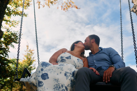 Couple sitting on a swing, sharing a kiss with a natural landscape in the background. Perfect for travel, romance, lifestyle, and adventure themes.の写真素材