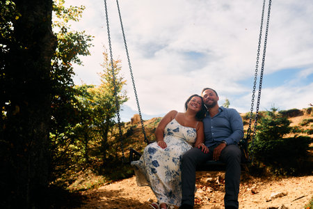 Couple sitting on a swing in the mountains, sharing a kiss with stunning natural landscape in the background. Perfect for travel, romance, lifestyle, and adventure themes.の写真素材