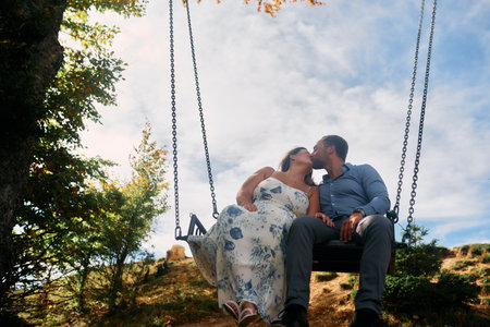 Couple sitting on a swing in the mountains, sharing a kiss with stunning natural landscape in the...