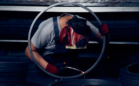 Skilled factory worker welding metal parts while wearing protective mask, gloves, and safety gear in...