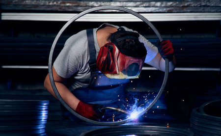 Skilled factory worker welding metal parts while wearing protective mask, gloves, and safety gear in...