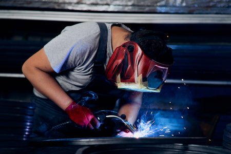 Skilled factory worker welding metal parts while wearing protective mask, gloves, and safety gear in...