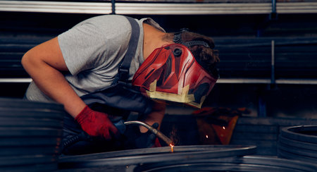 Skilled factory worker welding metal parts while wearing protective mask, gloves, and safety gear in...