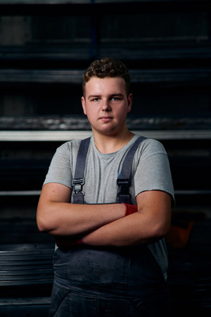 Confident young factory worker in grey tshirt and overalls standing with folded arms in industrial...