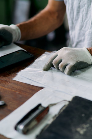 Close up of workers hands in white protective gloves reviewing technical documents and notes on wooden desk in industrial environment, symbolizing planning, organization, and quality control.の写真素材