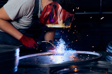 Skilled factory worker welding metal parts while wearing protective mask, gloves, and safety gear in...