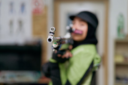 Female athlete wearing professional shooting uniform and hijab carefully aiming with an air rifle at an indoor range.の写真素材