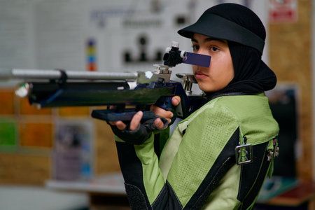 Female athlete wearing professional shooting uniform and hijab carefully aiming with an air rifle at an indoor range.の写真素材