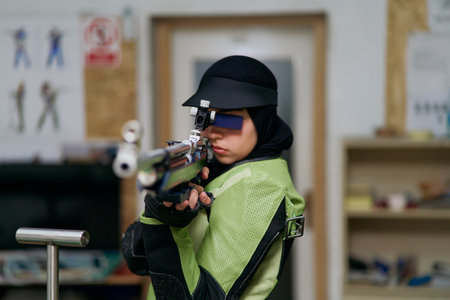 Female athlete wearing professional shooting uniform and hijab carefully aiming with an air rifle at an indoor range.の写真素材