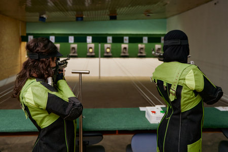 Two women practicing shooting at an indoor range, aiming rifles at targets, competitive sport,...