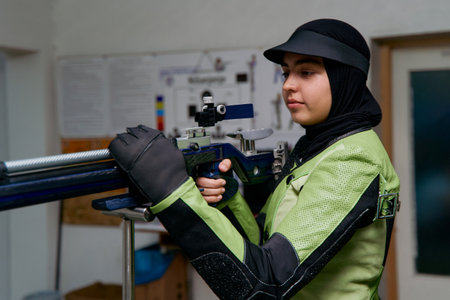 Focused female shooter in hijab aims rifle at target, indoor shooting range, competitive sport,...