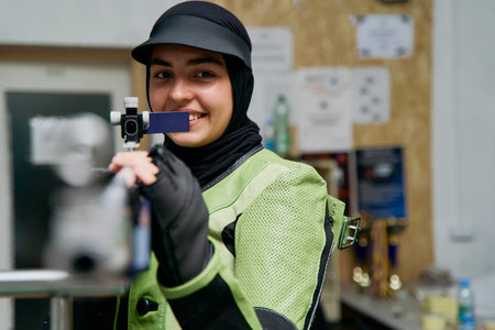 Smiling Muslim woman in hijab aiming with air rifle, indoor shooting range, competitive sport, focus...