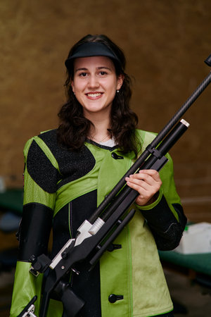 Smiling young woman holding a rifle, wearing shooting jacket and visor, ready for target practice,...