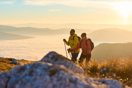 Couple hiking through a grassy mountain trail during sunrise, surrounded by a stunning backdrop of fog in the valleys below. The warm light creates a beautiful atmosphereの写真素材