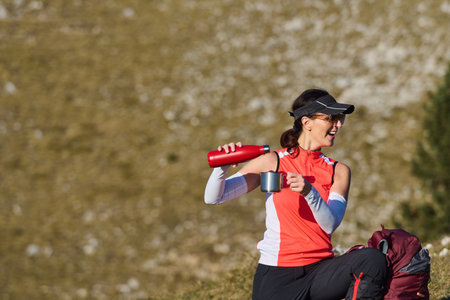 Woman enjoys a warm drink while resting on a mountain hike during sunny weatherの写真素材