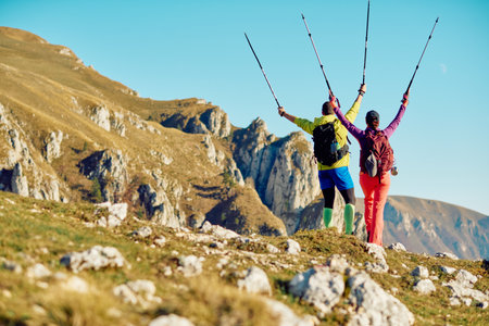 Two hikers celebrate their achievement with trekking poles on a mountain trail during a sunny day in natureの写真素材