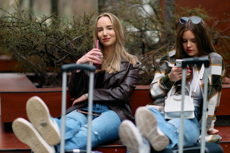 Two women sit on the city square with suitcases, two female tourists sit and check their mobile phones on the city square, a couple of female tourists explore the cityの写真素材