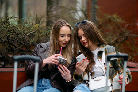 Two women sit on the city square with suitcases, two female tourists sit and check their mobile phones on the city square, a couple of female tourists explore the cityの写真素材