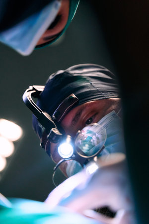 Surgeon In Headlamp Performing Operation Under Bright Surgical Lights In Modern OR During Critical Procedure With Teamの写真素材