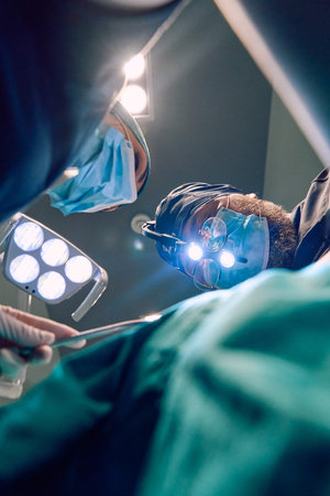 Surgeons In Operating Room Performing Surgery Under Bright Lights With Magnifying Loupes And Masks