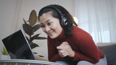 Asian woman on redshirt using a tablet with headphone for meeting online at home in pandemic...