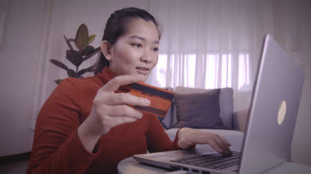 Asian women on redshirt using the laptop and credit card for shopping online in pandemic situation with a new normal lifestyleの写真素材