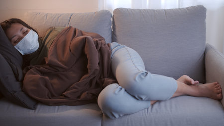Young Asian woman suffering from coronavirus has black eyes chills all over her body gets cold and uses a mask and blanket to keep warm on the sofa in her separate living room and quarantineの写真素材