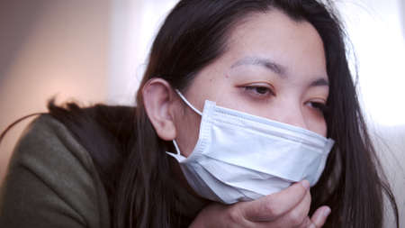 Young Asian woman suffering from coronavirus has black eyes chills all over her body gets cold and uses a mask and blanket to keep warm on the sofa in her separate living room and quarantineの写真素材
