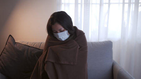 Young Asian woman suffering from coronavirus has black eyes chills all over her body gets cold and uses a mask and blanket to keep warm on the sofa in her separate living room and quarantineの写真素材