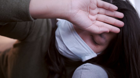 Young Asian woman suffering from coronavirus has black eyes chills all over her body gets cold and uses a mask and blanket to keep warm on the sofa in her separate living room and quarantineの写真素材