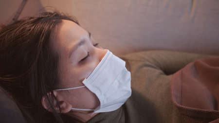 Young Asian woman suffering from coronavirus has black eyes chills all over her body gets cold and uses a mask and blanket to keep warm on the sofa in her separate living room and quarantineの写真素材