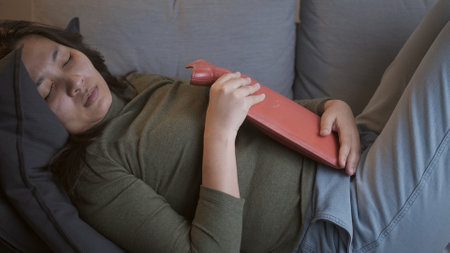 Sick woman with heavy menstrual cramps is applying a hot water bag on her stomach to relieve the...