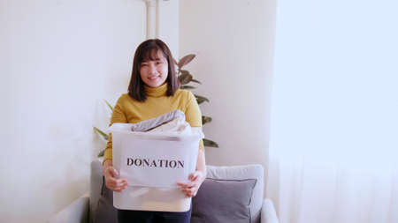 Portrait of young woman looking at camera holding box with clothes for donation. Concept campaign to...